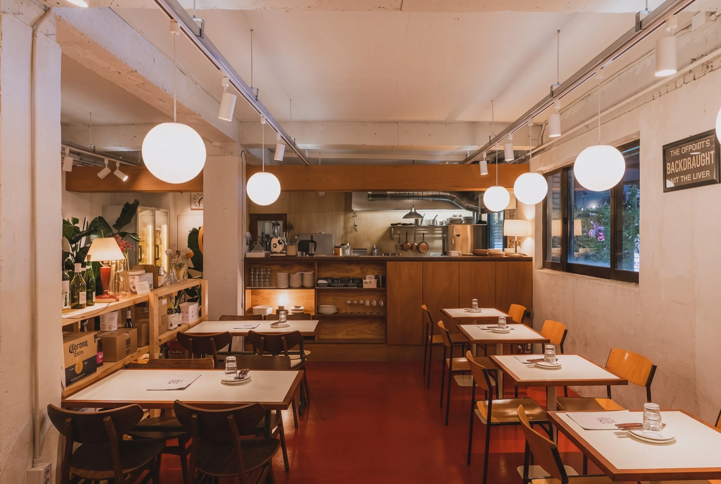 The counter runs the length of the room with woven baskets and wine bottles on the left shelf, an open kitchen with copper pans hanging under a range hood, wood tables with dark and light chairs on a red floor, and round white globe pendants hanging from track lighting along the ceiling
